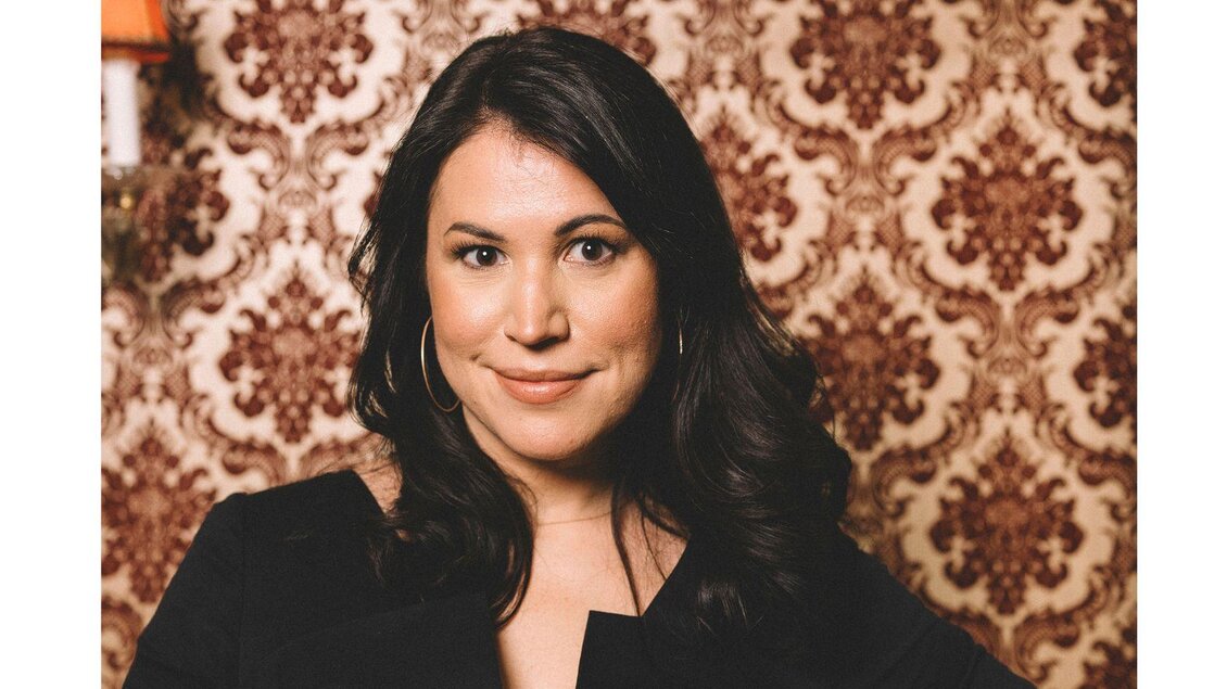 Eine Frau mit langen, dunklen Haaren lächelt in die Kamera. Im Hintergrund ist eine klassische, gemusterte Wandtapete zu sehen. | © Die Wasnerin