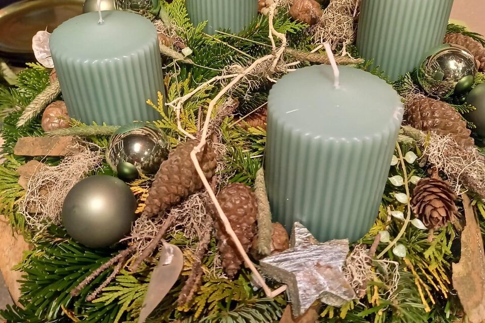 Ein schön dekorierter Adventskranz mit vier blauen Kerzen, umgeben von Tannenzweigen, Zapfen und glitzernden Ornamenten. Die Anordnung strahlt eine gemütliche und festliche Atmosphäre aus. | © Blumenhof Bender