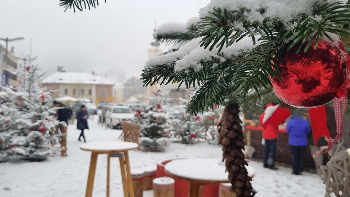 Ein verschneiter Weihnachtsmarkt mit Tannenbäumen und festlicher Dekoration. Menschen schlendern durch die winterliche Atmosphäre. | © Stadt Mürzzuschlag, Bettina Rinnhofer