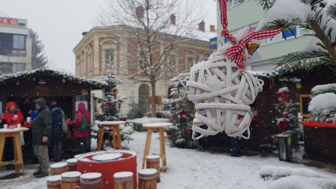 Ein festlich dekorierter Weihnachtsmarkt im Schnee. Eine weiße Weihnachtsbaumdekoration hängt an einem Baum, während Besucher in warmen Kleidern die Atmosphäre genießen. | ©  Stadt Mürzzuschlag, Bettina Rinnhofer