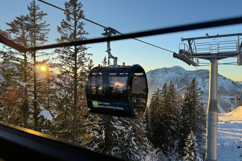 Eine Gondelbahn fährt durch eine verschneite Landschaft mit hohen Tannen. Im Hintergrund sind Berge und ein klarer Himmel bei Sonnenaufgang zu sehen. | © Theresa Schwaiger_TVB Ausseerland Salzkammergut