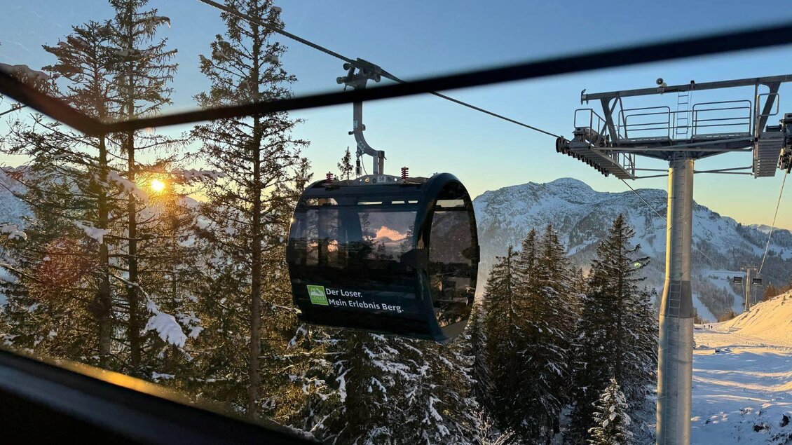 Eine Gondelbahn fährt durch eine verschneite Landschaft mit hohen Tannen. Im Hintergrund sind Berge und ein klarer Himmel bei Sonnenaufgang zu sehen. | © Theresa Schwaiger_TVB Ausseerland Salzkammergut