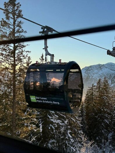 A gondola ride travels through a snowy landscape with tall fir trees. In the background, there are mountains and a clear sky at sunrise. | © Theresa Schwaiger_TVB Ausseerland Salzkammergut