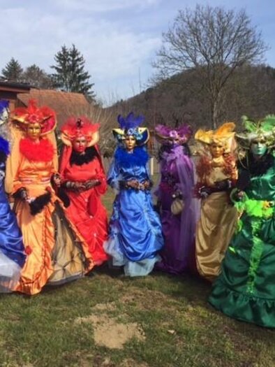 Eine Gruppe von Personen in farbenfrohen Kostümen und Masken steht im Freien. Der Hintergrund zeigt eine ruhige Landschaft mit Bäumen und Wiesen. | © Anni Rathkolb