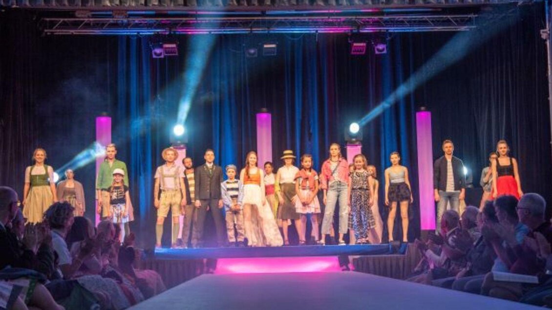 Eine Modenschau mit mehreren Models auf dem Laufsteg. Im Hintergrund sind bunte Lichter und ein applaudierendes Publikum zu sehen. | © www.narzissenfest.at/Sams