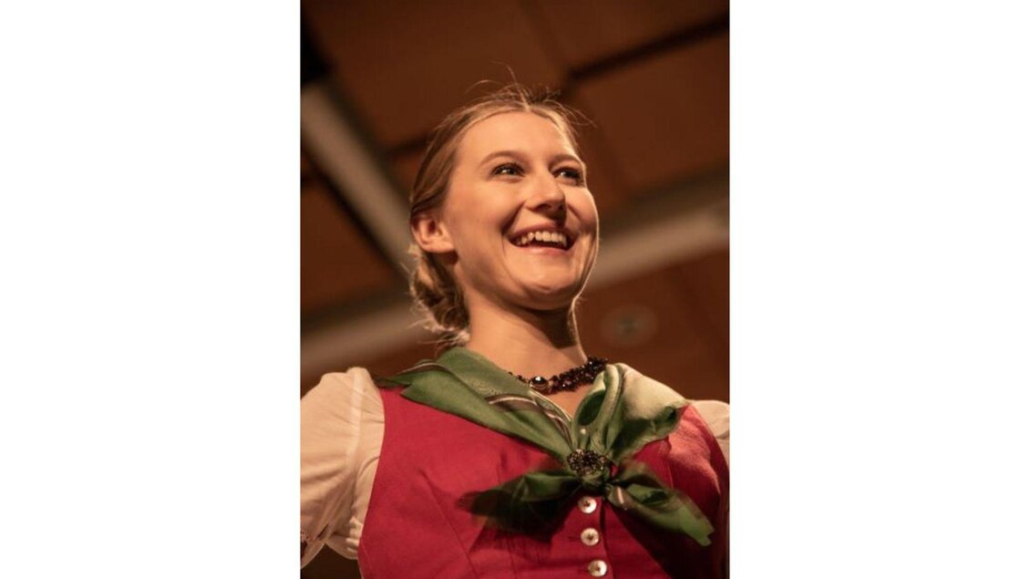 Eine fröhliche Frau trägt ein traditionelles Outfit mit einer roten Bluse und einer grünen Schleife. Sie lächelt und wirkt gut gelaunt. | © www.narzissenfest.at/Sams