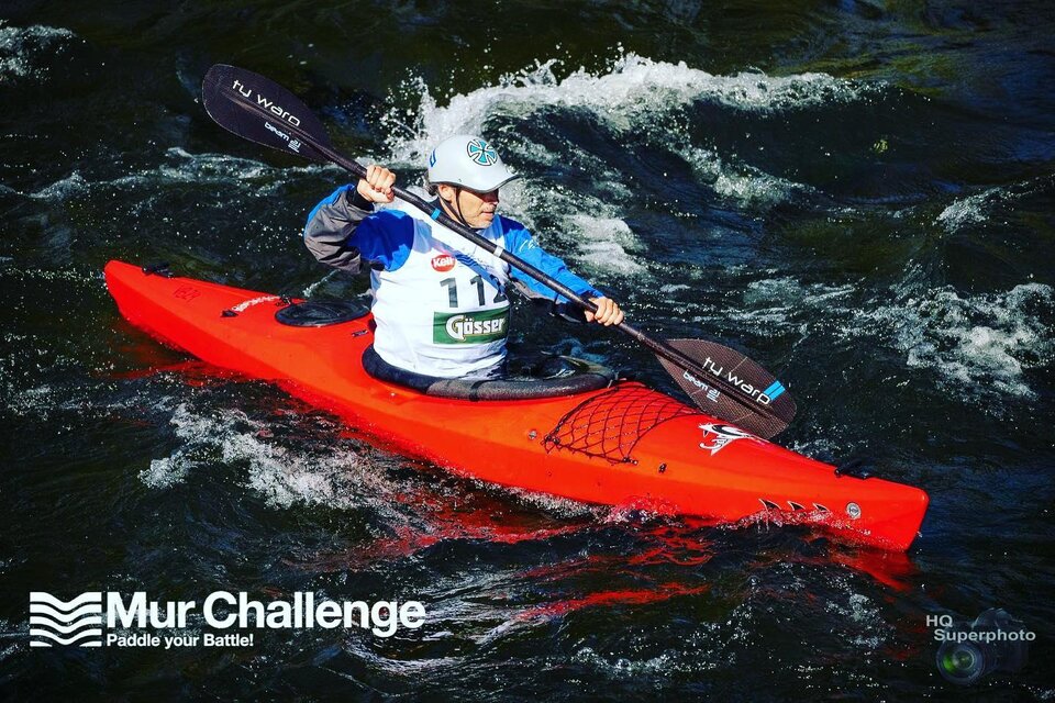Ein Kanute paddelt in einem roten Kajak auf einem reißenden Fluss. Spritzwasser und Wellen umgeben ihn in einer dynamischen Wettkampfszene. | © HQ Superphoto
