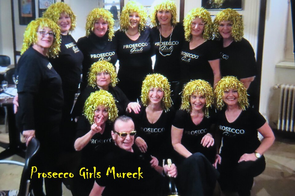 Eine Gruppe von Frauen mit gelben Lockenperücken steht in schwarzer Kleidung zusammen. Sie lächeln fröhlich und halten eine Flasche Prosecco. | © Peter Raffler