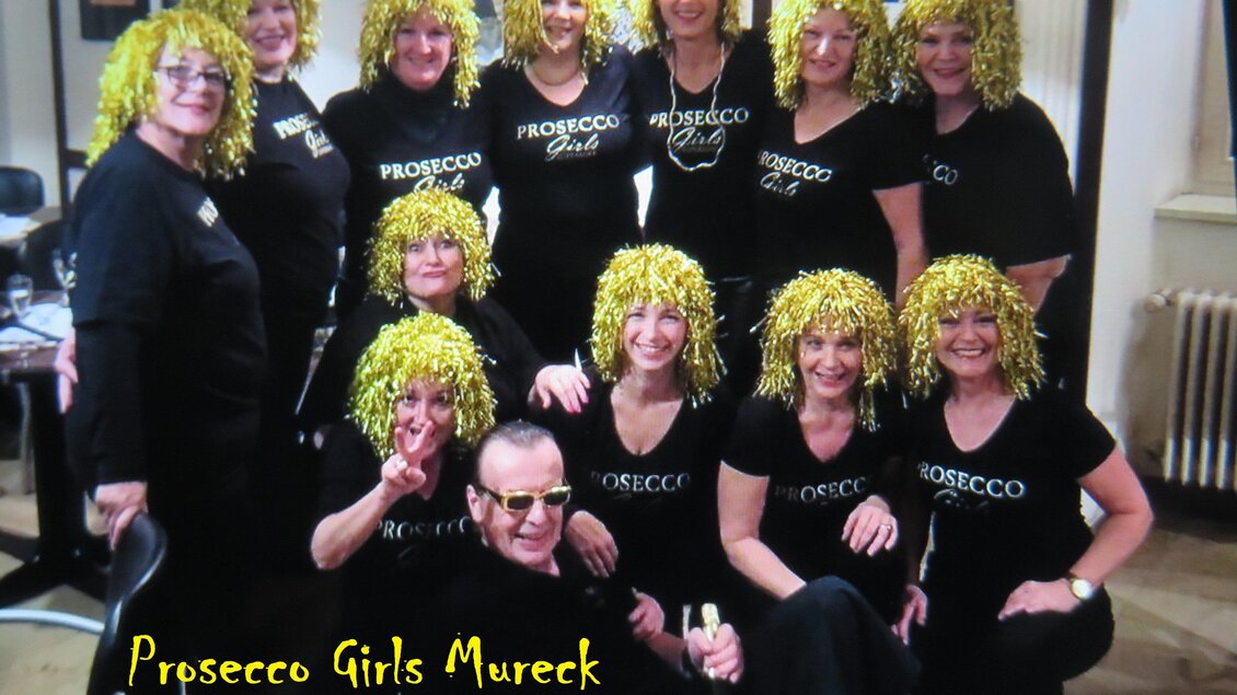 Eine Gruppe von Frauen mit gelben Lockenperücken steht in schwarzer Kleidung zusammen. Sie lächeln fröhlich und halten eine Flasche Prosecco. | © Peter Raffler