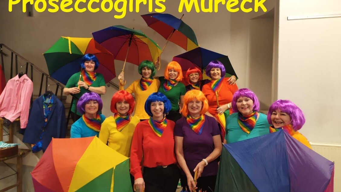 Eine Gruppe von fröhlichen Frauen in bunten Perücken steht zusammen und hält Regenschirme in Regenbogenfarben. Sie tragen bunte Kleidung und strahlen Spaß und Lebensfreude aus. | © Peter Raffler