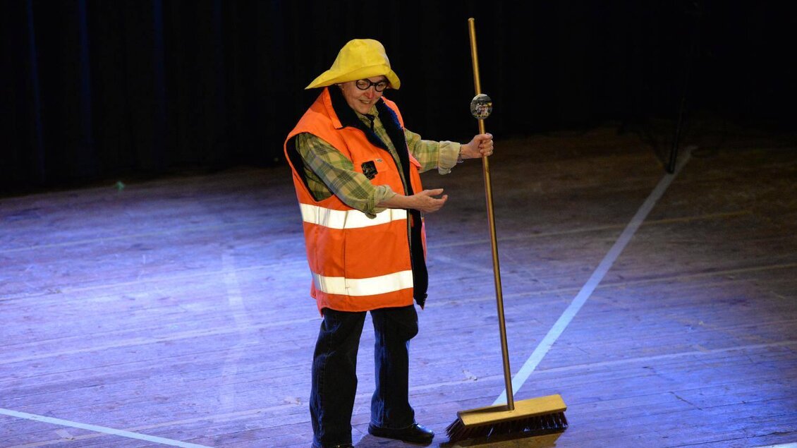 Ein Mann trägt eine leuchtend orangefarbene Warnweste und einen gelben Hut. Er hält einen Besen und steht auf einer Bühne. | © Werner Friedl