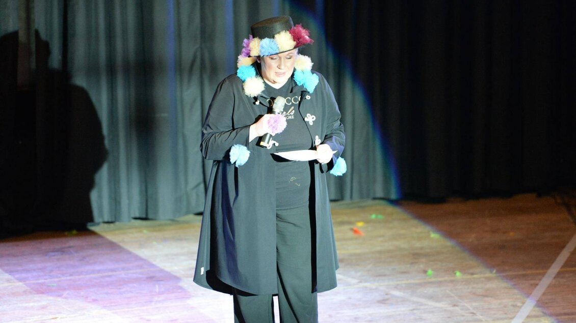 Eine Person auf der Bühne trägt ein Kostüm mit bunten Accessoires. Sie hält ein Mikrofon und scheint eine Ansprache zu halten. | © Werner Friedl