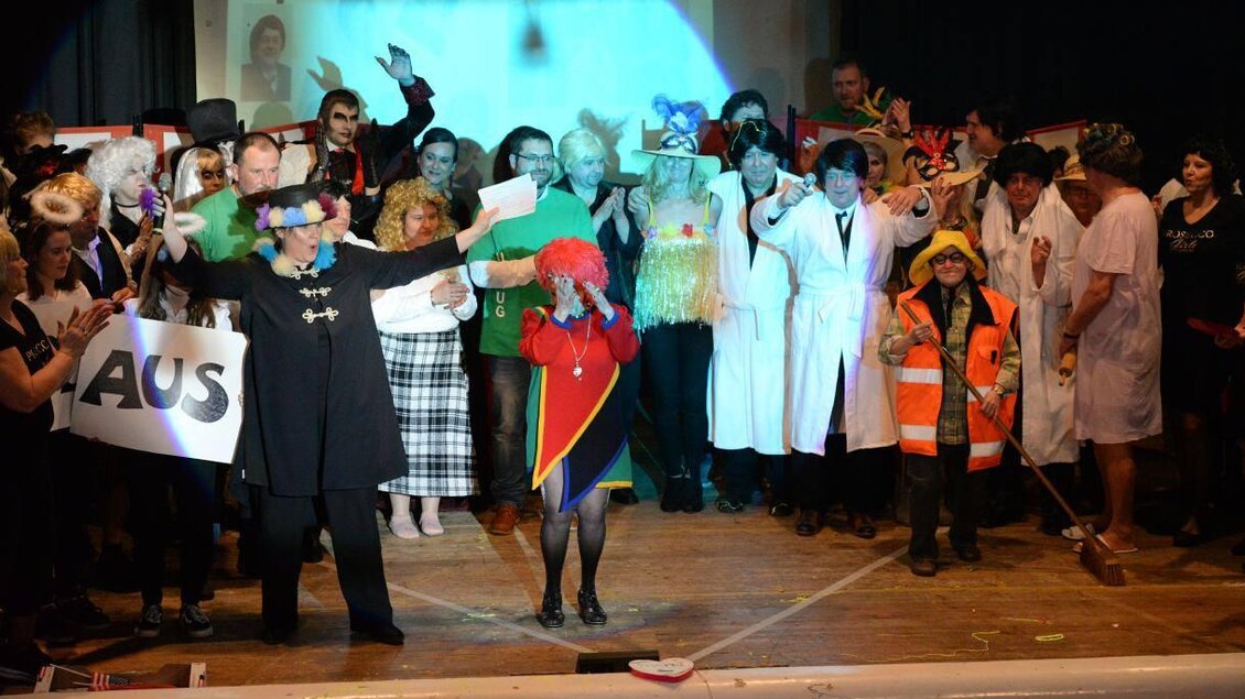 Eine bunte Gruppe von Menschen auf einer Bühne, die verschiedene Kostüme und Requisiten tragen. Einige halten Schilder, während sie feiern und posieren. | © Werner Friedl