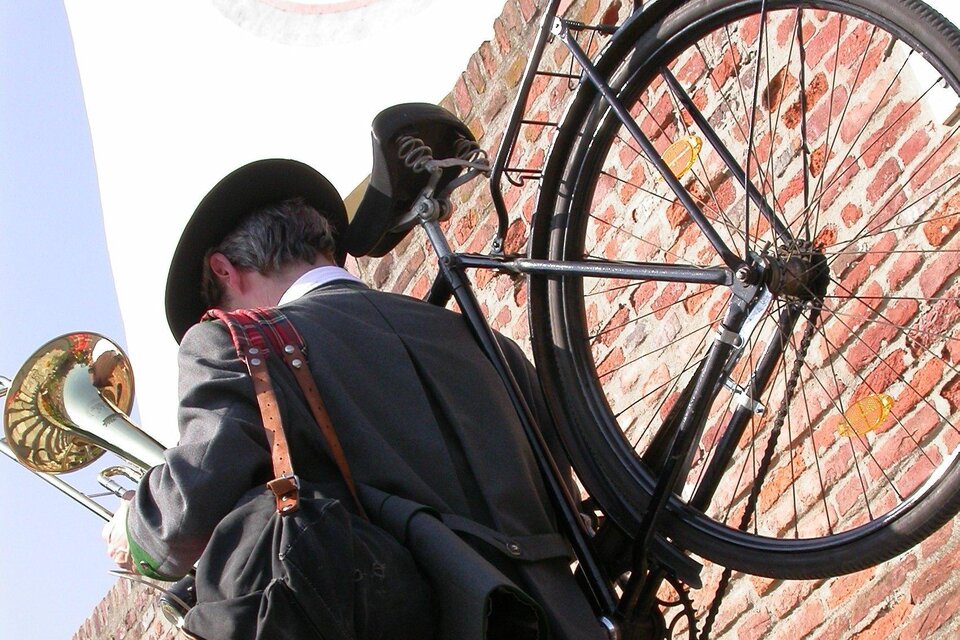 Ein Musiker in traditioneller Kleidung trägt ein Blasinstrument und eine Tasche, während er mit einem Fahrrad durch die Straßen von Graz geht. Der Musiker ist von einer historischen Backsteinmauer umgeben, was auf die kulturelle Atmosphäre eines musikalischen Stadtspaziergangs hinweist. | © STVLW
