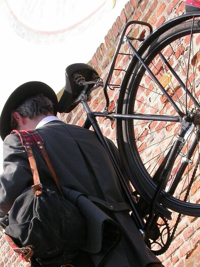 Ein Musiker in traditioneller Kleidung trägt ein Blasinstrument und eine Tasche, während er mit einem Fahrrad durch die Straßen von Graz geht. Der Musiker ist von einer historischen Backsteinmauer umgeben, was auf die kulturelle Atmosphäre eines musikalischen Stadtspaziergangs hinweist. | © STVLW