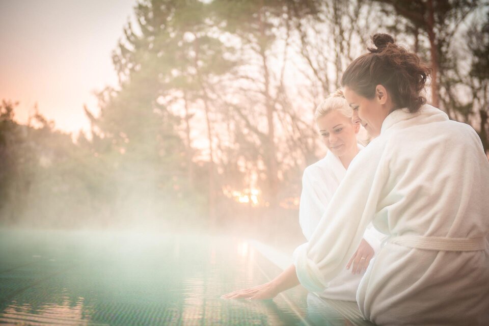 Zwei Frauen in Bademänteln entspannen am Rand eines Schwimmbeckens. Im Hintergrund sind Bäume und ein sanfter Sonnenuntergang zu sehen. | © RETTER Bio- Natur- Resort