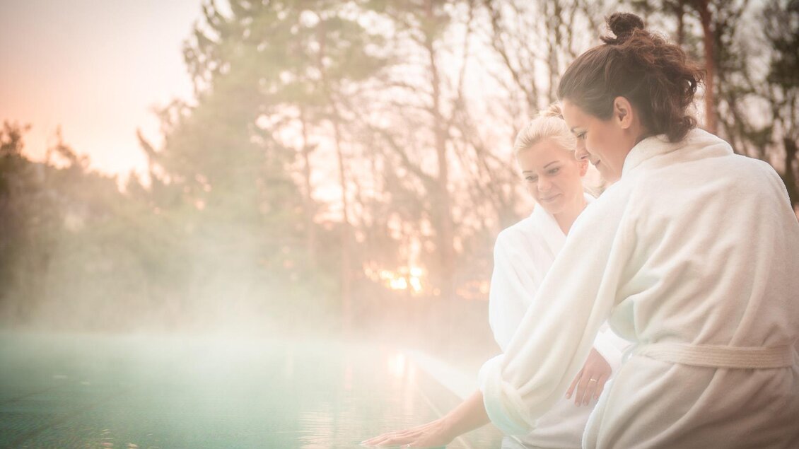 Zwei Frauen in Bademänteln entspannen am Rand eines Schwimmbeckens. Im Hintergrund sind Bäume und ein sanfter Sonnenuntergang zu sehen. | © RETTER Bio- Natur- Resort