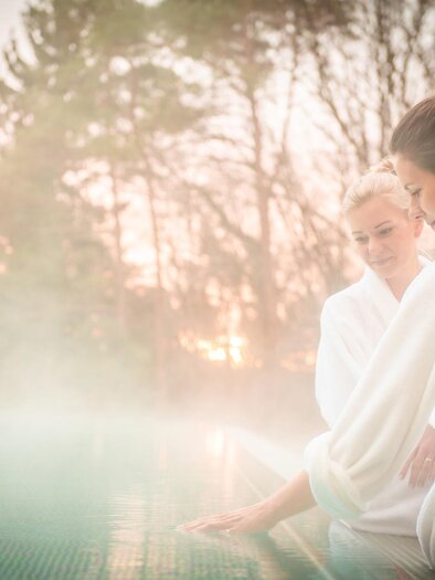 Two women in bathrobes relax at the edge of a swimming pool. In the background, trees and a gentle sunset are visible. | © RETTER Bio- Natur- Resort