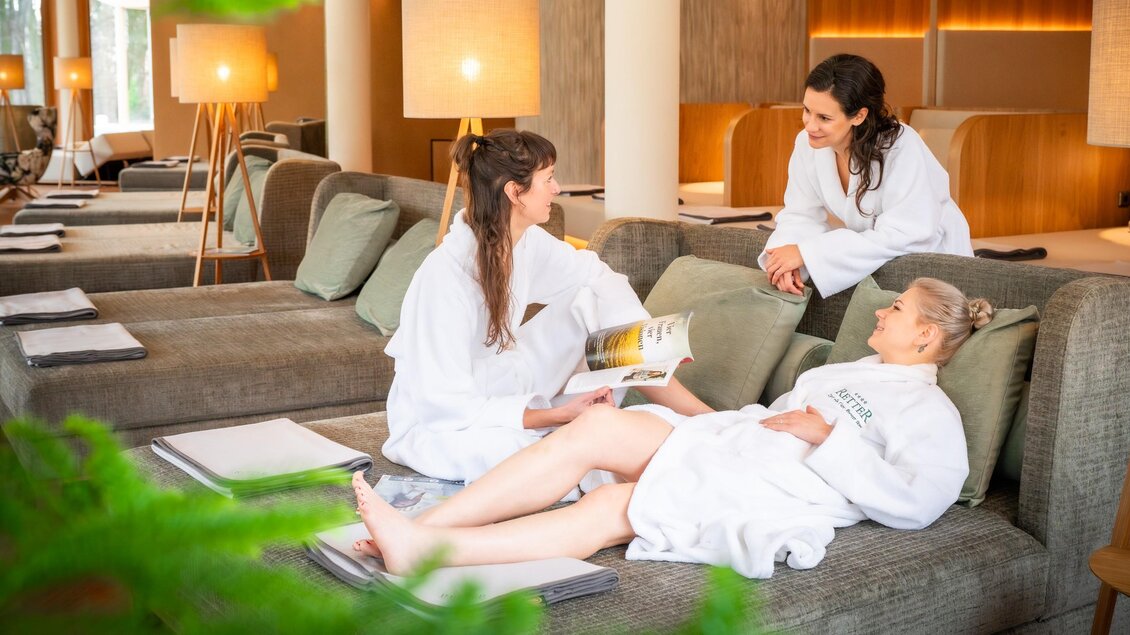 Drei Frauen in Bademänteln entspannen in einem modernen Spa. Eine Frau liest in einem Magazin, während die anderen beiden entspannt im Loungebereich sitzen. | © RETTER Bio- Natur- Resort