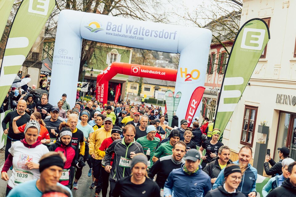 Eine Gruppe von Läufern startet bei einem Rennen in Bad Waltersdorf. Bunte Sportbekleidung und ein großer Startbogen prägen die Szene. | © Hikimus