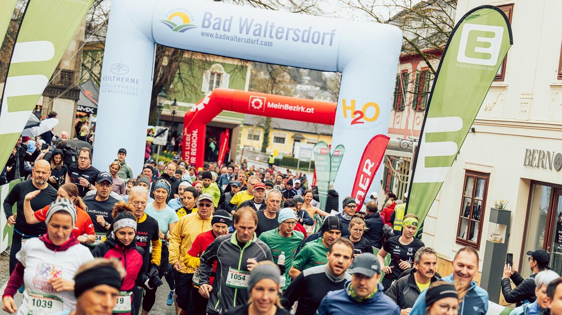 Eine Gruppe von Läufern startet bei einem Rennen in Bad Waltersdorf. Bunte Sportbekleidung und ein großer Startbogen prägen die Szene. | © Hikimus