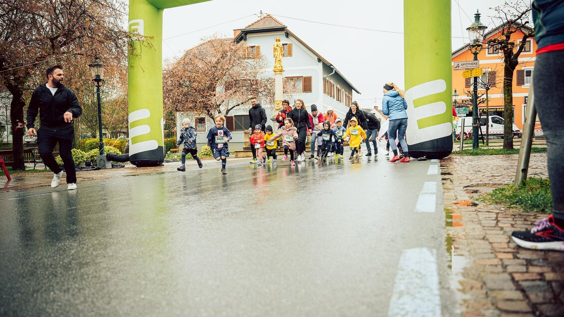 Eine Gruppe von Kindern und Erwachsenen läuft unter einem großen grünen Startbogen. Die Straße ist nass, und die Umgebung ist städtisch mit Häusern und Bäumen. | © Hikimus