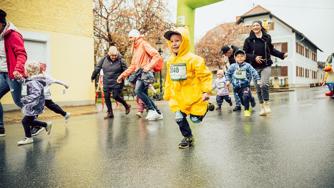 Eine Gruppe von Kindern läuft fröhlich auf der Straße, während es leicht regnet. Einige Erwachsene begleiten sie, und im Hintergrund ist ein Startbogen zu sehen. | © Hikimus