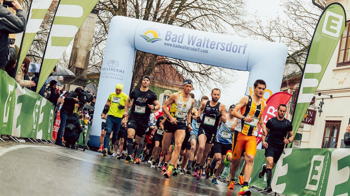 Ein Wettlauf mit mehreren Läufern, die unter einem Startbogen in Bad Waltersdorf hindurchlaufen. Die Strecke ist nass und es sind zahlreiche Zuschauer anwesend. | © Hikumus