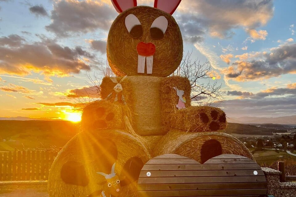 Ein großer Heuballen-Hase mit roten Ohren steht vor einem malerischen Sonnenuntergang. Im Vordergrund befindet sich eine gemütliche Bank.