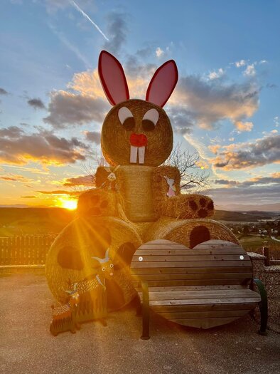A large hay bale bunny with red ears stands in front of a picturesque sunset. In the foreground, there is a cozy bench.