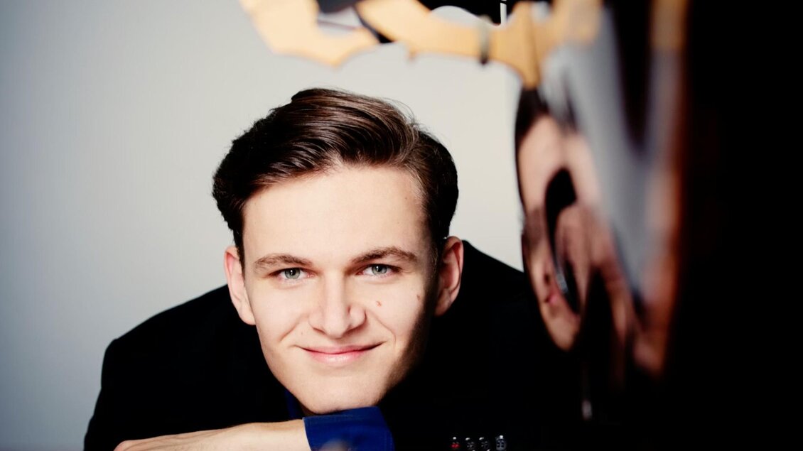 Ein Porträt von Dominik Wagner, einem jungen Mann, der entspannt auf einem Kontrabass sitzt. Er trägt einen schwarzen Anzug mit einem blauen Hemd und lächelt leicht in die Kamera. Der Hintergrund ist unscharf, wodurch der Fokus auf seinem Gesicht und der eleganten Haltung liegt. Der Kontrabass ist teilweise im Bild sichtbar, und es ist ein kreativer, professioneller Ausdruck zu erkennen. | © Julia Wesely