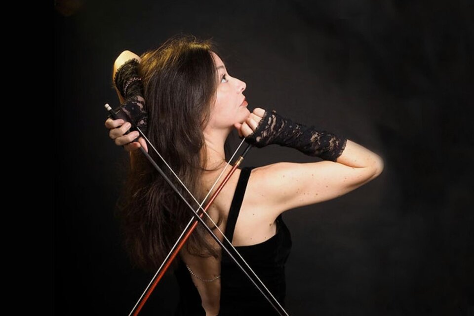 Lina Tur Bunet, eine Geigerin und Dirigentin, posiert in einem eleganten schwarzen Kleid, während sie zwei Geigenbögen in der Hand hält. Sie ist von der Seite fotografiert, mit einem nach oben gerichteten Blick und einem ausdrucksstarken, konzentrierten Gesicht. Ihre Hände sind in schwarze Spitzenhandschuhe gehüllt, und der dunkle Hintergrund verstärkt die dramatische Atmosphäre des Bildes. | © Juarez
