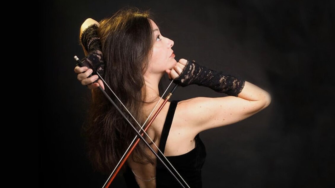 Lina Tur Bunet, eine Geigerin und Dirigentin, posiert in einem eleganten schwarzen Kleid, während sie zwei Geigenbögen in der Hand hält. Sie ist von der Seite fotografiert, mit einem nach oben gerichteten Blick und einem ausdrucksstarken, konzentrierten Gesicht. Ihre Hände sind in schwarze Spitzenhandschuhe gehüllt, und der dunkle Hintergrund verstärkt die dramatische Atmosphäre des Bildes. | © Juarez