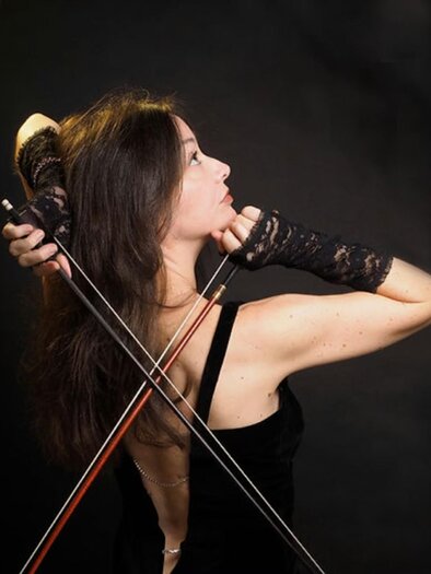 Lina Tur Bunet, a violinist and conductor, poses in an elegant black dress while holding two violin bows in her hands. She is photographed from the side, with an upward gaze and an expressive, focused face. Her hands are enveloped in black lace gloves, and the dark background enhances the dramatic atmosphere of the image. | © Juarez