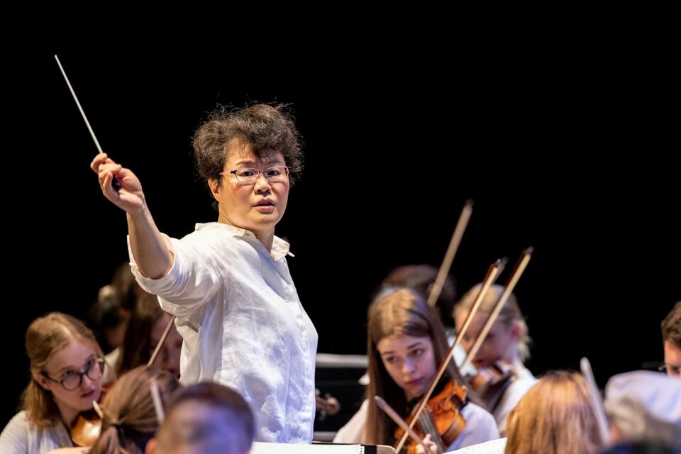 Dirigentin Mei-Ann Chen leitet das Orchester bei einem Konzert in Graz während der Veranstaltung "Recreation". Sie steht auf der Bühne, hält den Taktstock in der Luft und dirigiert mit Ausdruck und Energie. Im Hintergrund sind Orchestermusiker zu sehen, die ihre Instrumente spielen, während die Atmosphäre des Konzerts intensiv und fokussiert ist. | © Nikola Milatovic