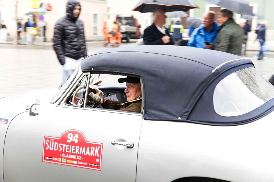 Ein klassisches Auto fährt durch eine belebte Straße. Im Fahrzeug sitzt ein älterer Mann mit einem Lächeln. | © Fotostudio Semrad