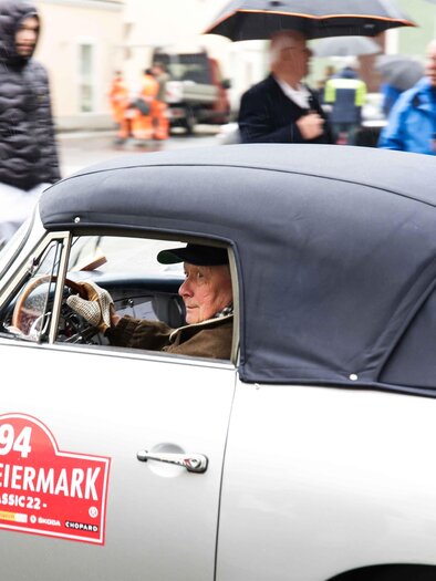 A classic car drives through a bustling street. Inside the vehicle sits an older man with a smile. | © Fotostudio Semrad