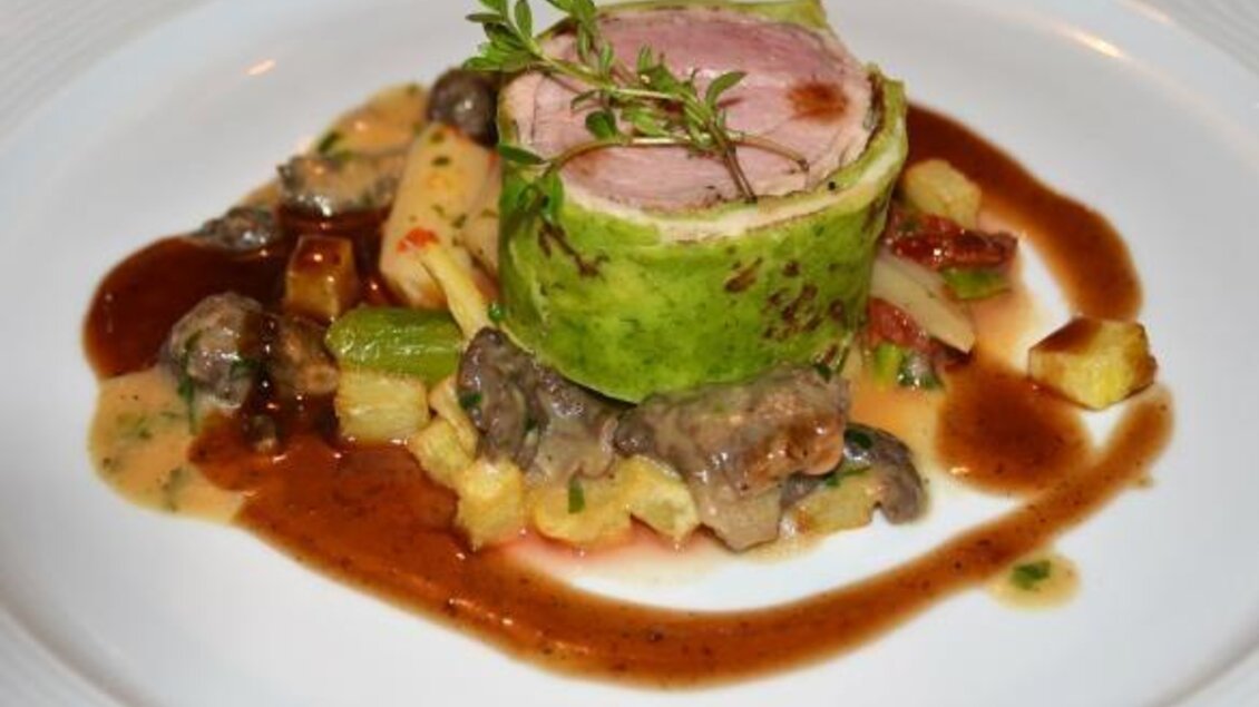 Ein kunstvoll angerichtetes Gericht mit einer grünen Gemüse-Rolle und Fleisch. Am Rand sind verschiedene Beilagen und Saucen platziert.