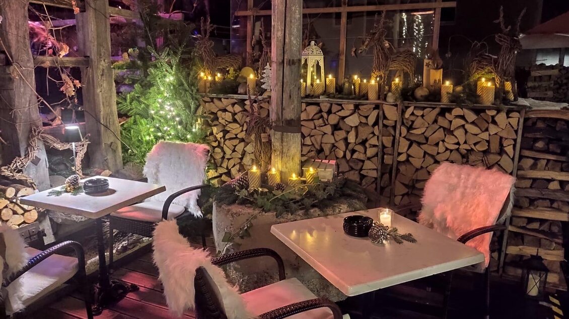 Eine gemütliche Außenterrasse mit zwei Tischen und weichen, weißen Decken. Die Atmosphäre wird durch Kerzenlicht und Holzscheite im Hintergrund erwärmt. | © Stocker Peter