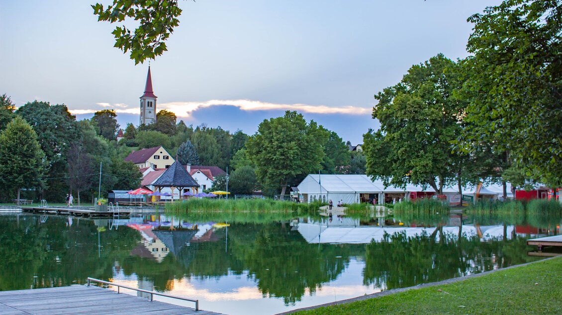 Ein ruhiger See mit spiegelndem Wasser und umgeben von Bäumen. Im Hintergrund sind Häuser und ein Kirchturm sichtbar. | © Sandra Brünner