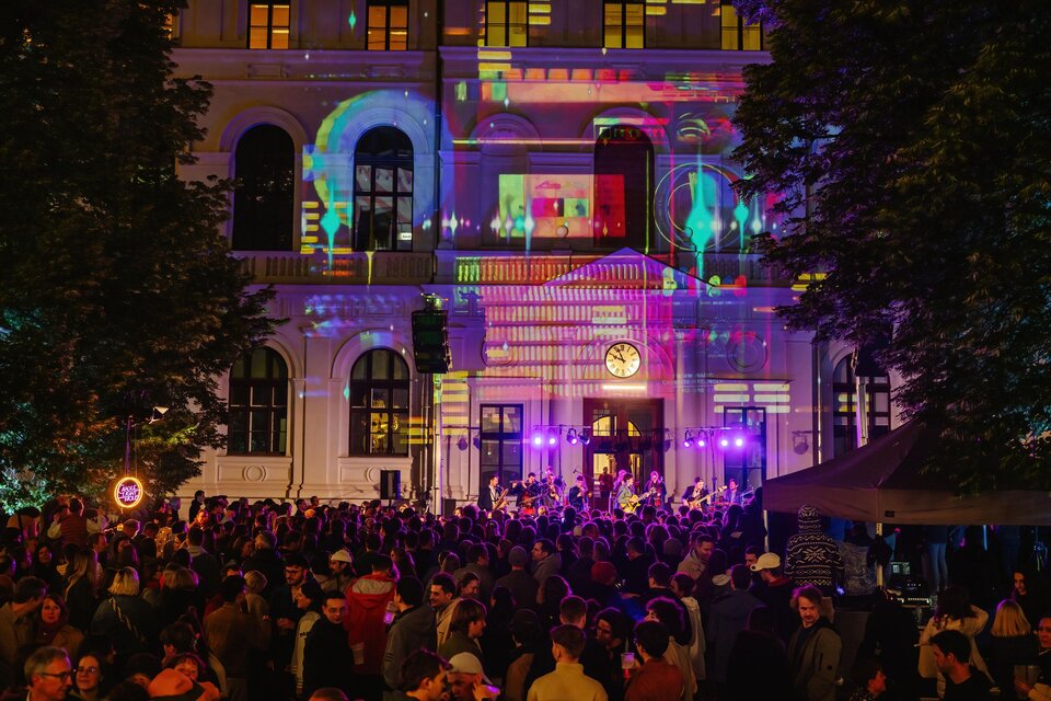 Eine große Menschenmenge bei einem Abend-Event vor einem beleuchteten Gebäude. Die Fassade ist mit bunten Lichtern und Projektionen geschmückt. | © Nadja_Fuchs_Uni Vibes