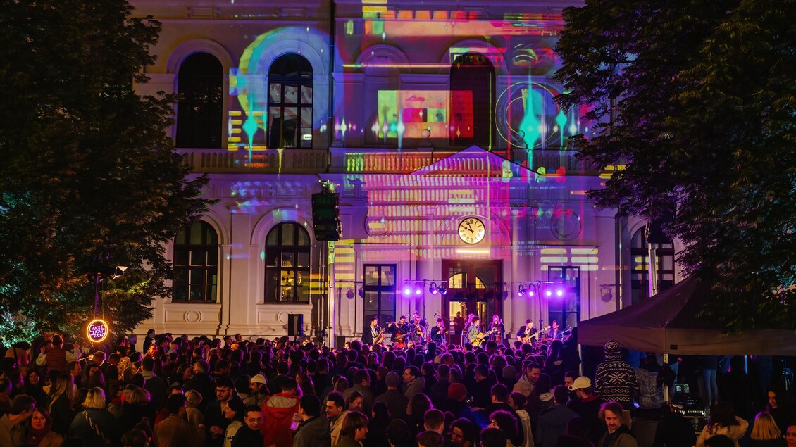 Eine große Menschenmenge bei einem Abend-Event vor einem beleuchteten Gebäude. Die Fassade ist mit bunten Lichtern und Projektionen geschmückt. | © Nadja_Fuchs_Uni Vibes