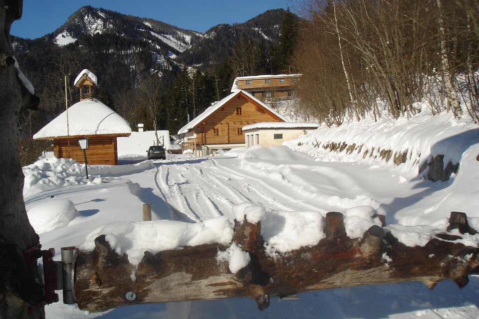 Eine verschneite Landschaft mit einem Holzhaus und einem kleinen Glockenturm. Die Spuren im Schnee führen zu den Gebäuden im Vordergrund, umgeben von Bergen. | © Hangler Huabn