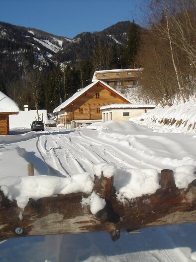 A snowy landscape with a wooden house and a small bell tower. The tracks in the snow lead to the buildings in the foreground, surrounded by mountains. | © Hangler Huabn