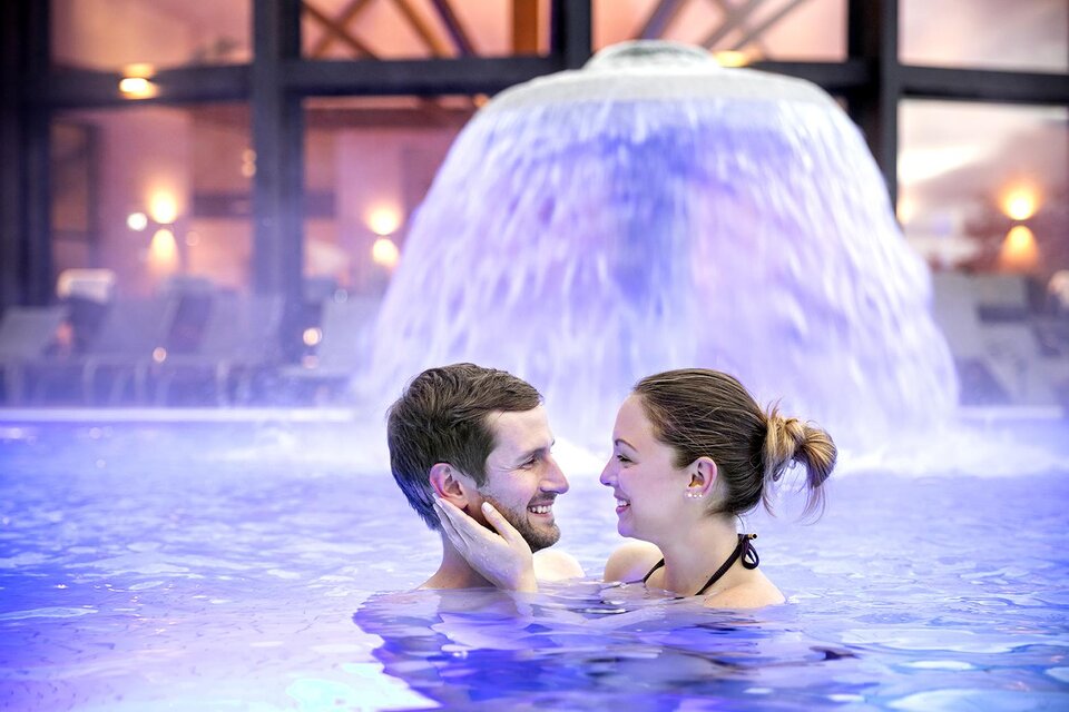 Ein Paar steht lächelnd im Wasser vor einem Wasserfall. Die Atmosphäre ist romantisch und entspannend. | © Eisenberger_@Heilthermen Resort Bad Waltersdorf