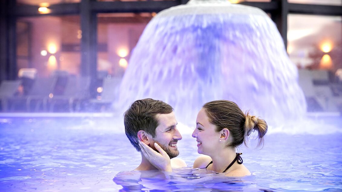 Ein Paar steht lächelnd im Wasser vor einem Wasserfall. Die Atmosphäre ist romantisch und entspannend. | © Eisenberger_@Heilthermen Resort Bad Waltersdorf