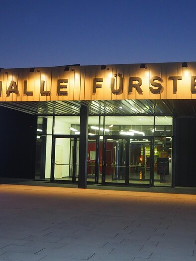 The town hall of Fürstenfeld is illuminated at dusk. It has modern architecture with large glass windows and striking light accents.
