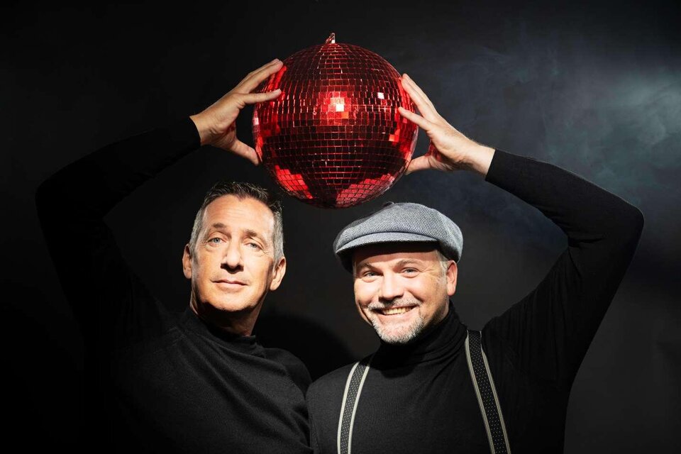 Zwei Männer stehen vor einem dunklen Hintergrund und halten eine rote Discokugel über ihren Köpfen. Sie tragen schwarze Oberteile und lächeln freundlich. | © LukasBeck