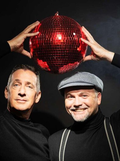 Two men stand in front of a dark background holding a red disco ball above their heads. They are wearing black tops and smiling kindly. | © LukasBeck