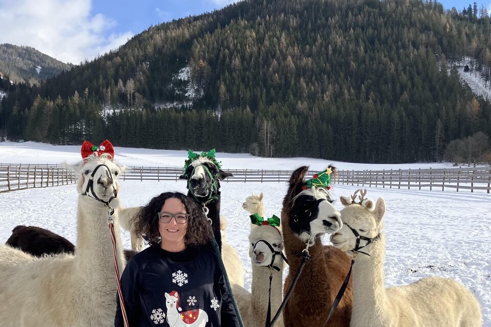 Eine Frau steht im Schnee mit mehreren Alpakas, die festliche Accessoires tragen. Im Hintergrund sind Wälder und Berge zu sehen. | © Alpaka, Lamas und Rentiere zum Grünen See